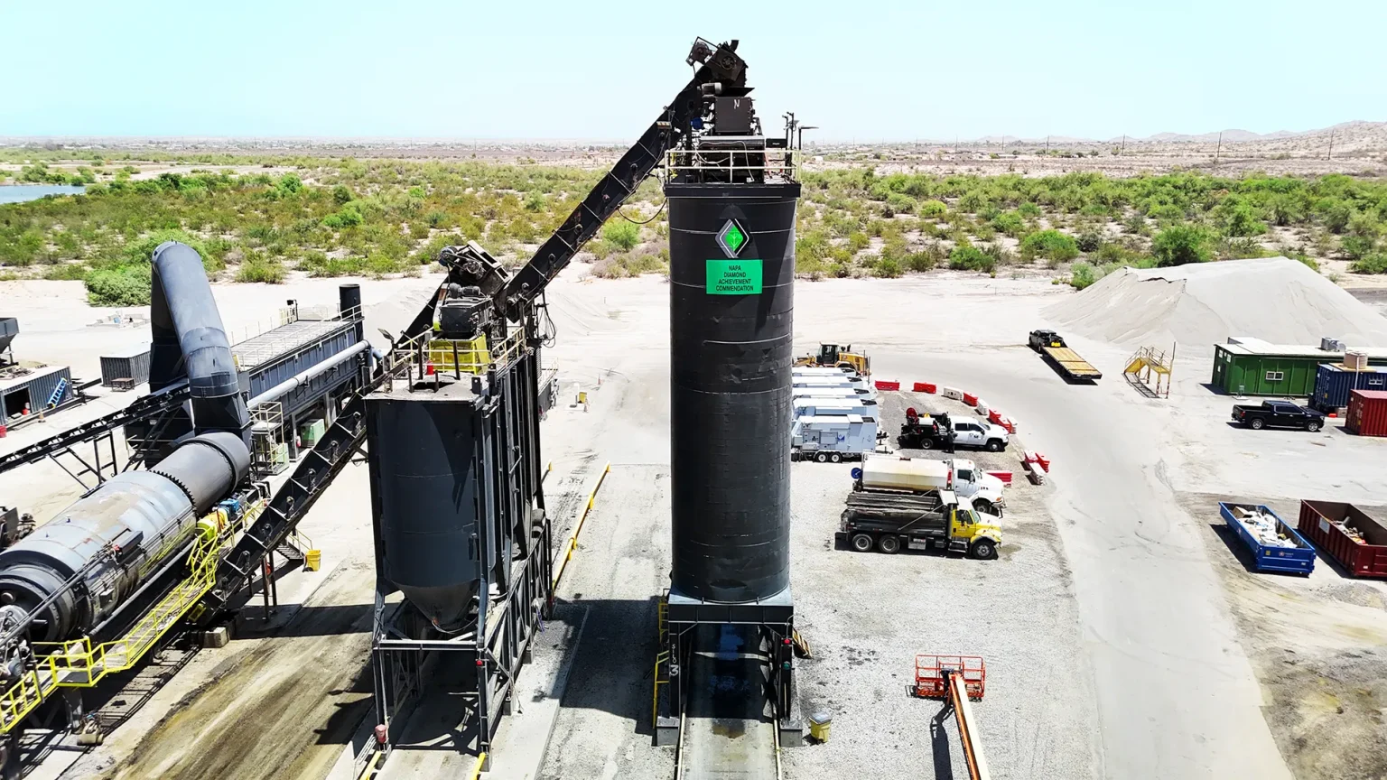 A large industrial silo at an outdoor construction site with machinery, trucks, and gravel piles under a clear sky.