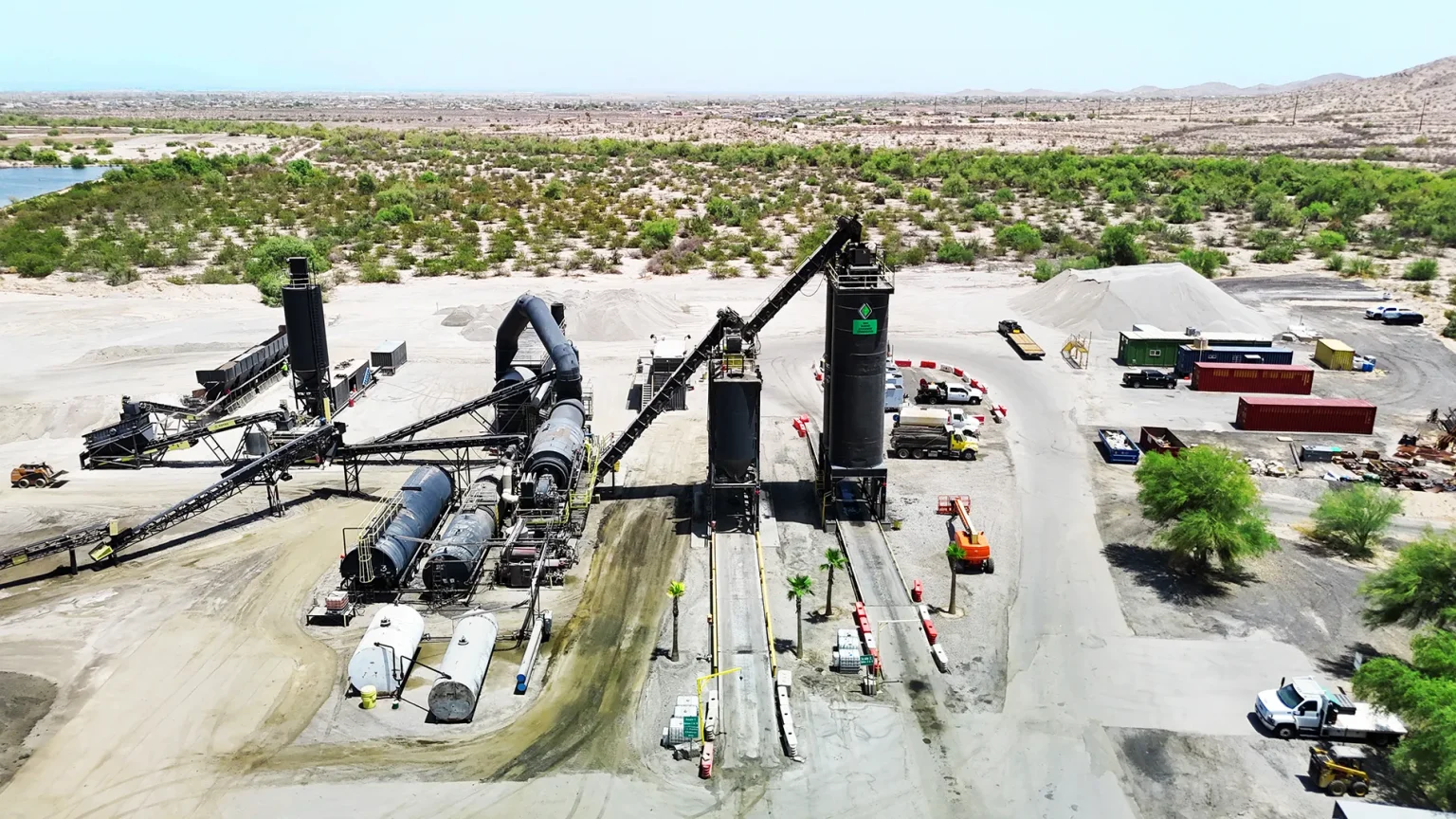Aerial view of an industrial asphalt mixing plant with machinery, storage tanks, and construction materials in a desert landscape.