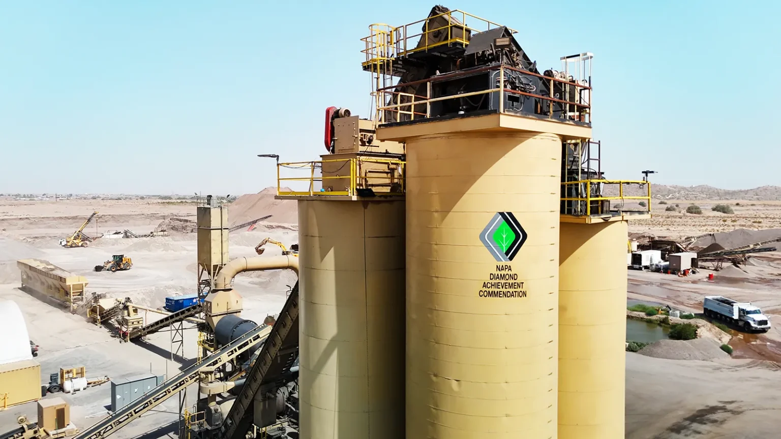 A large industrial facility with beige storage silos stands in a desert landscape, displaying a logo reading "NAPA Diamond Achievement Commendation.