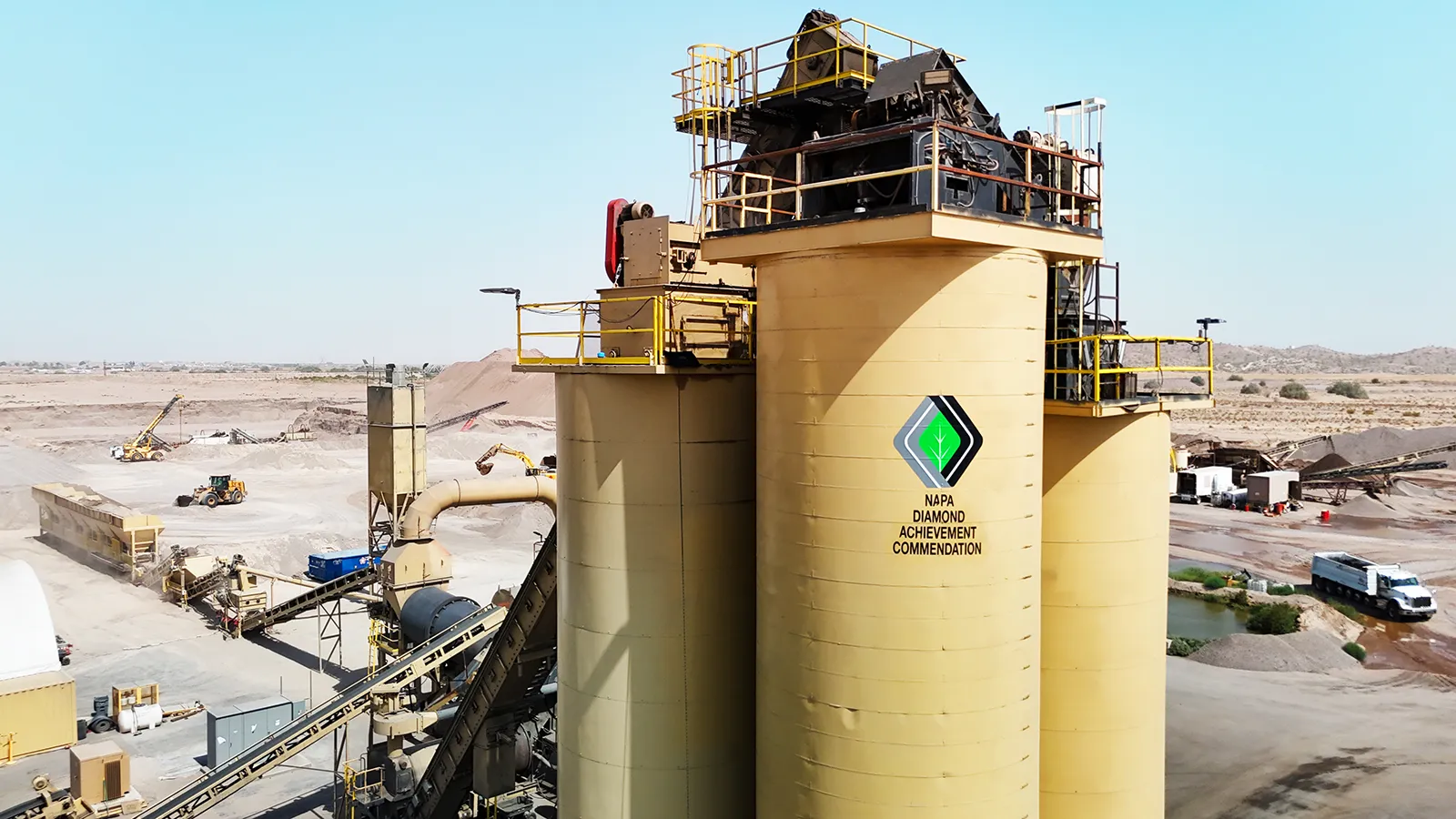 A large industrial facility with beige storage silos stands in a desert landscape, displaying a logo reading "NAPA Diamond Achievement Commendation.