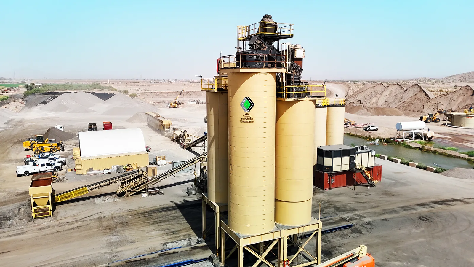 A large industrial facility with yellow storage silos, machinery, and construction vehicles set in a dry, open landscape.