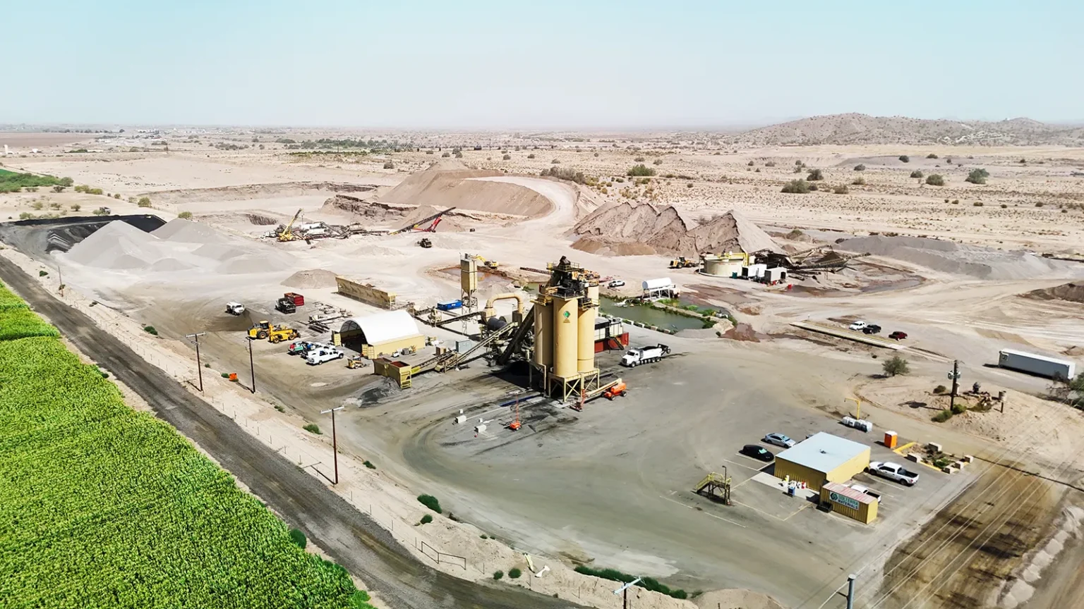 Aerial view of an industrial mining or processing facility with equipment, vehicles, and storage piles in a desert landscape near a green field.