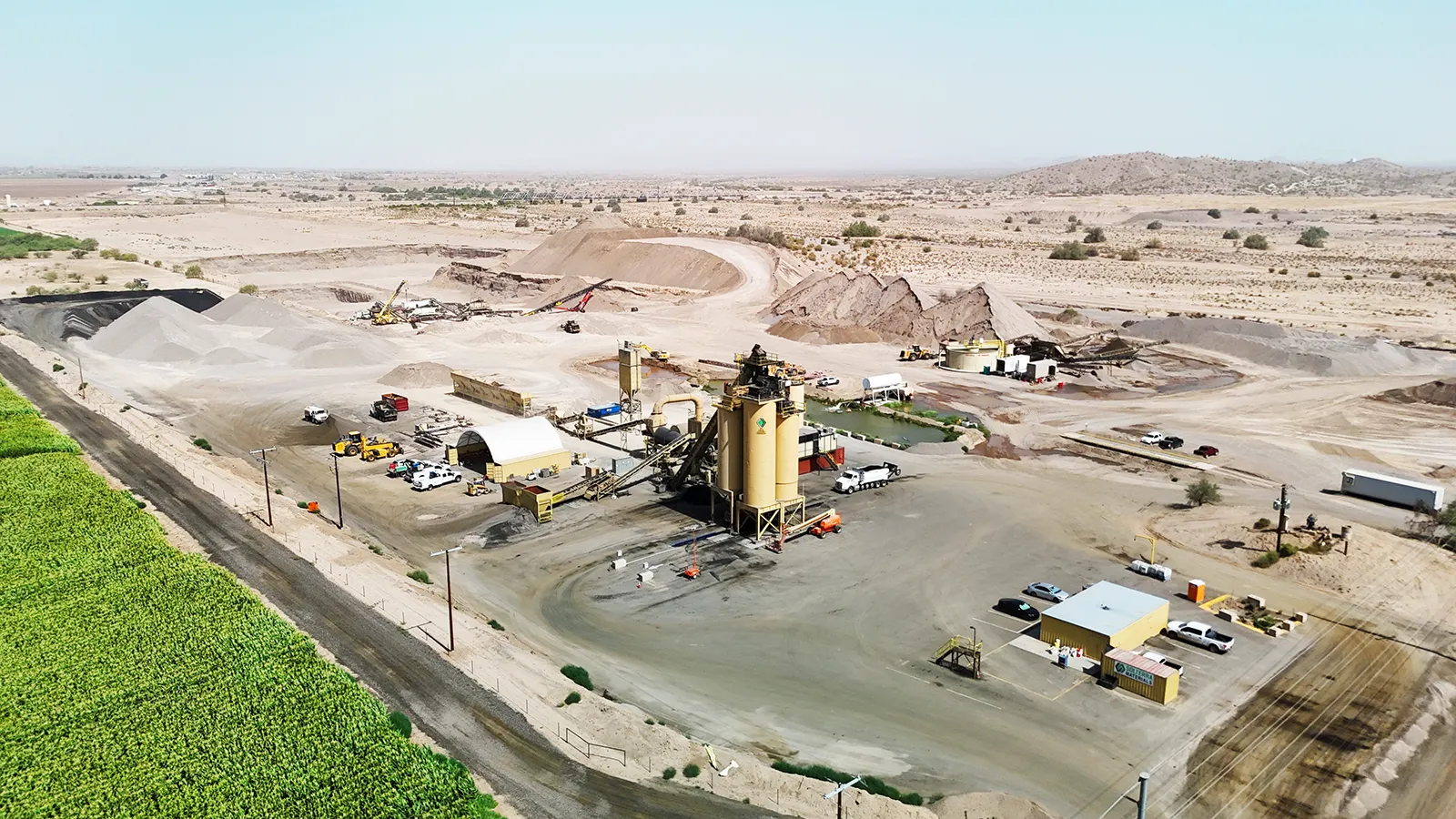 Aerial view of an industrial mining or processing facility with equipment, vehicles, and storage piles in a desert landscape near a green field.
