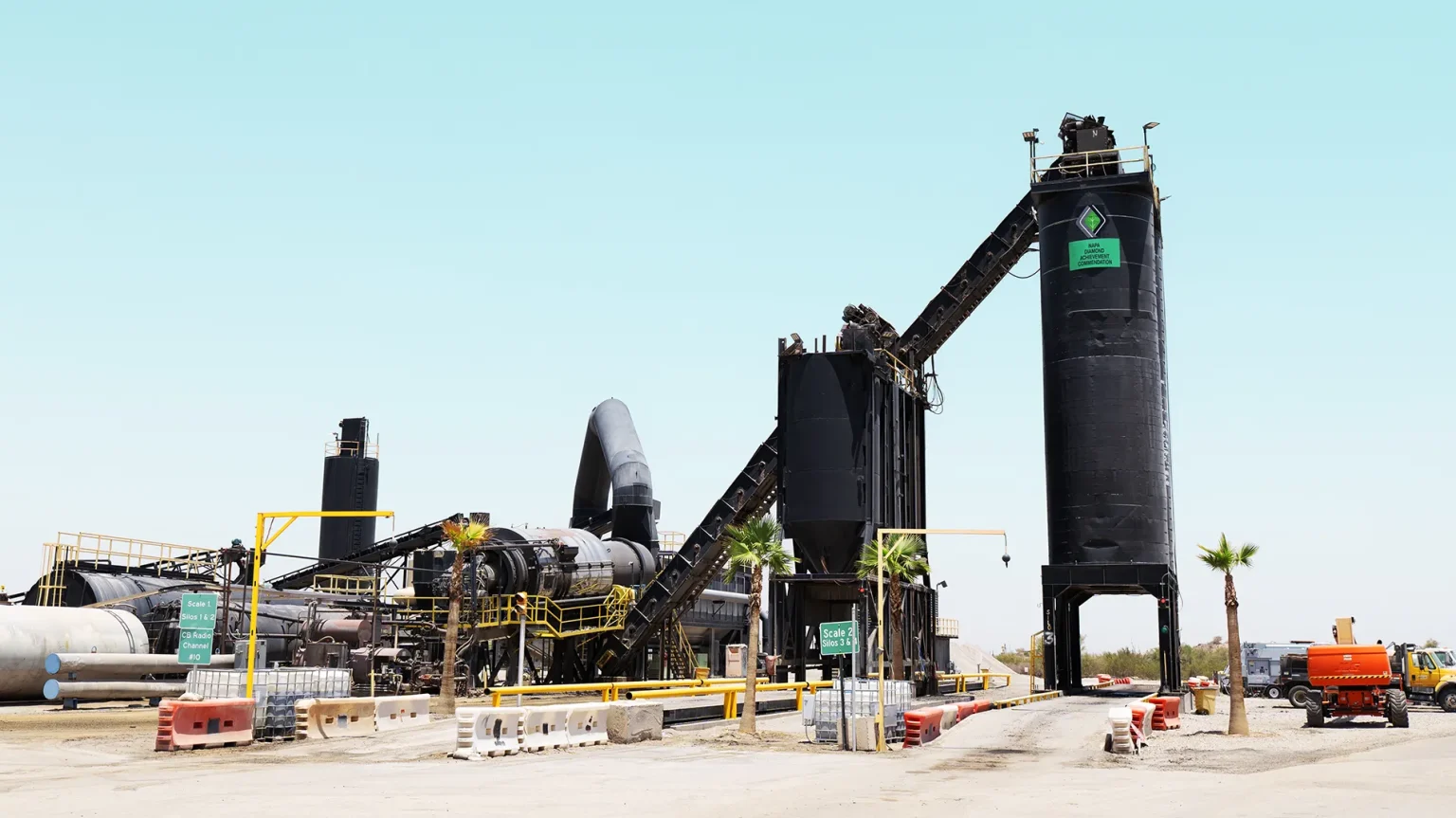 An industrial asphalt plant with large black silos, conveyor belts, and machinery, surrounded by barriers, palm trees, and clear blue sky in the background.