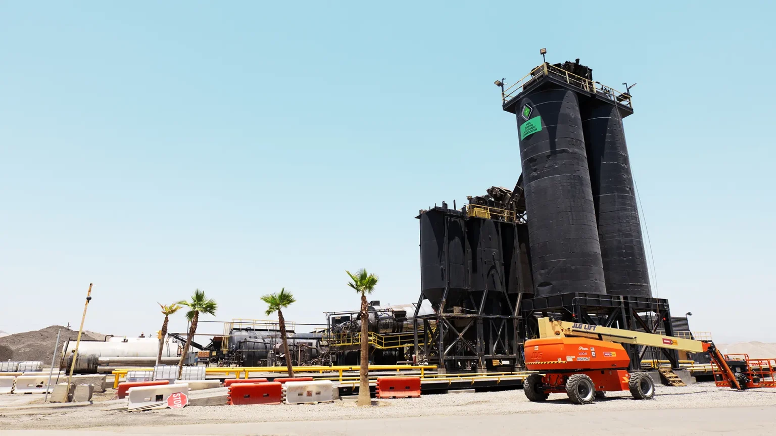 An industrial facility with two large black silos, safety barriers, construction equipment, and a few palm trees in front, under a clear blue sky.