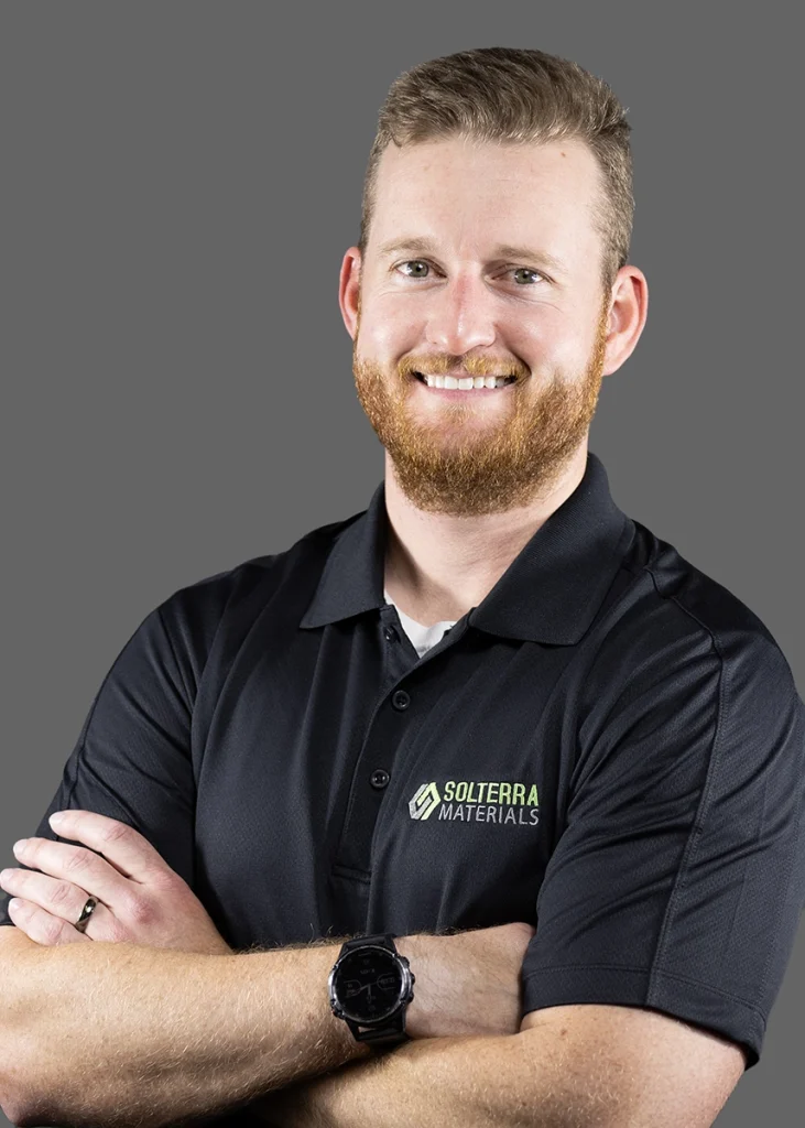 A man with a red beard wearing a black Solterra Materials polo shirt stands with arms crossed, smiling, in front of a plain gray background.