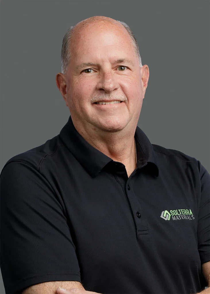 A middle-aged man with short hair and a mustache wearing a black Solterra Materials polo shirt stands against a gray background, smiling with arms crossed.