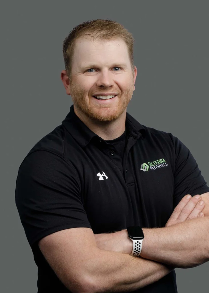 A man with light hair and a beard, wearing a black polo shirt with a Sittera Materials logo, stands against a gray background with arms crossed and a smartwatch on his wrist.