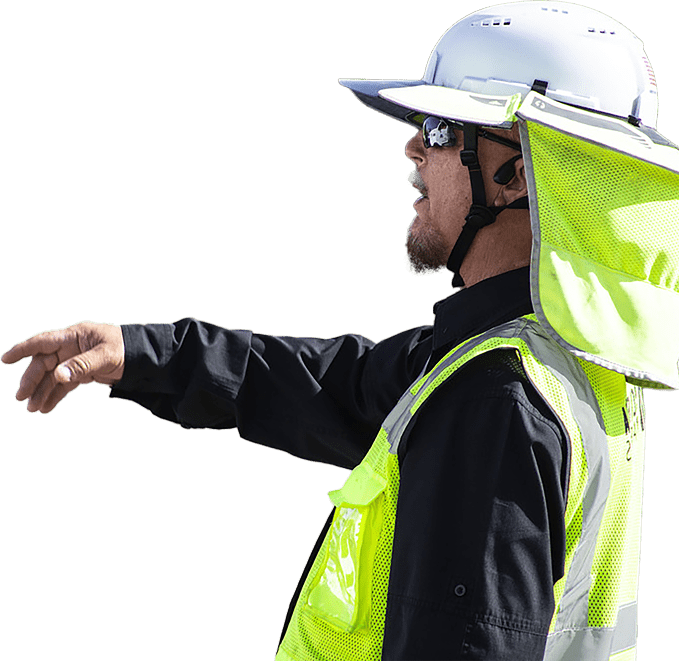 A construction worker in a safety vest, helmet, and sunglasses points with his right arm extended.