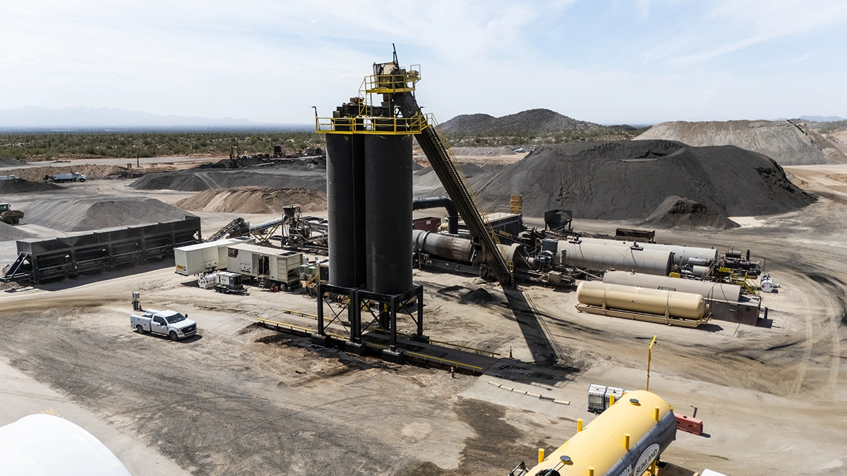 Aerial view of an industrial asphalt plant with storage tanks, conveyors, equipment, vehicles, and piles of raw materials surrounded by a dry landscape.