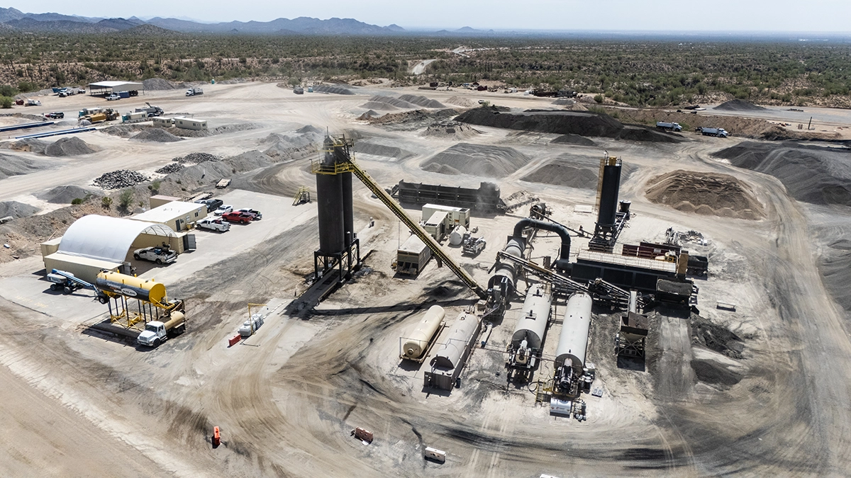 Aerial view of an industrial mining or processing facility with large machinery, storage tanks, and surrounding piles of raw materials in a desert landscape.