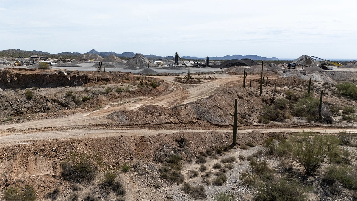 A dry, desert landscape with dirt roads, sparse vegetation, cacti, and piles of earth and rocks, with industrial equipment visible in the background.