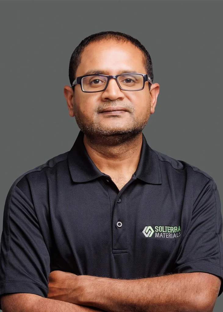 A man with short hair and glasses wearing a black Solterra Materials polo shirt stands with his arms crossed against a plain gray background.