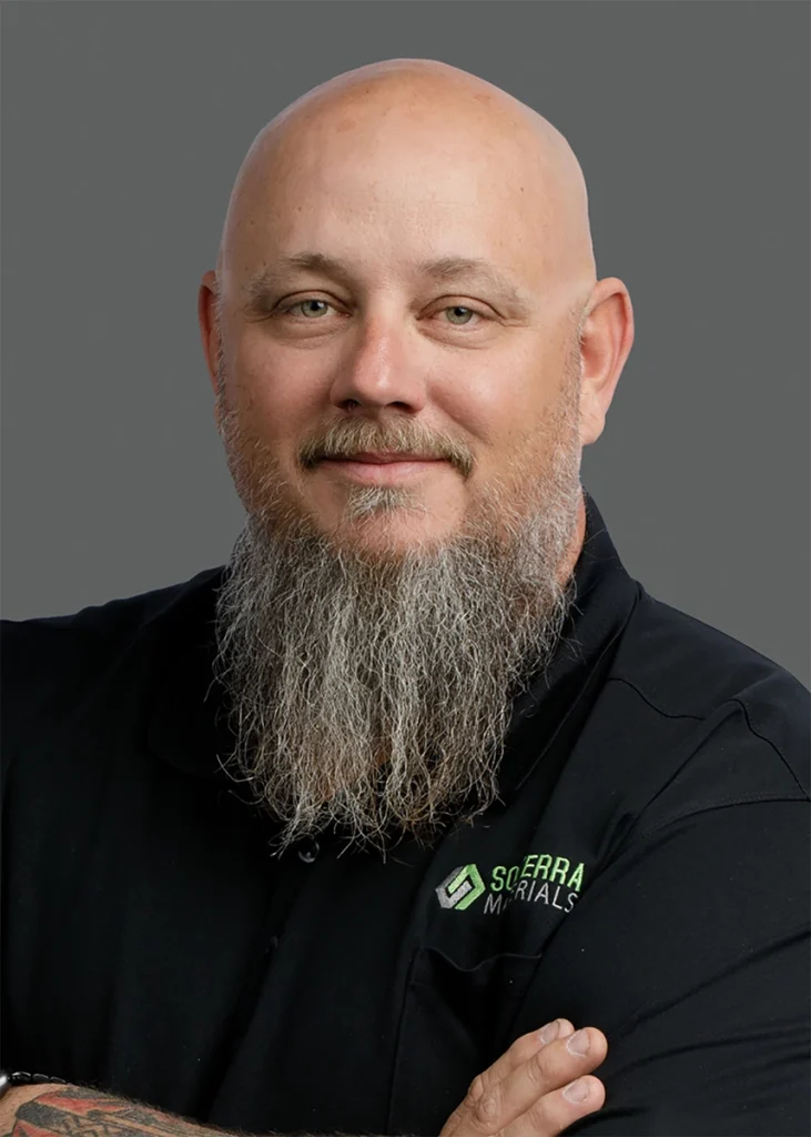 A bald man with a long gray beard wearing a black polo shirt with a "Sierra Materials" logo, standing against a plain gray background.
