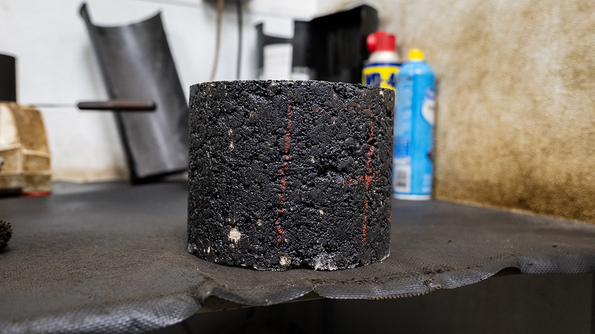 A cylindrical core sample of asphalt sits on a workbench in a workshop, with tools and spray cans visible in the background.