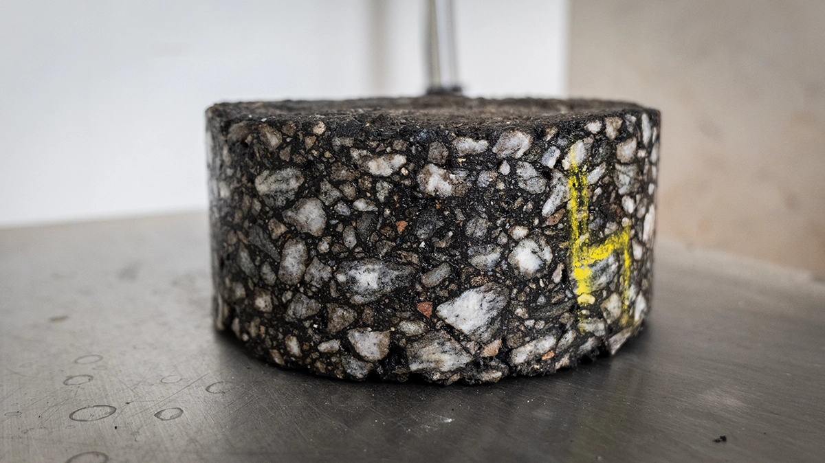 A cylindrical core sample of asphalt with visible gravel inclusions and a yellow marking on its side, placed on a metal surface.