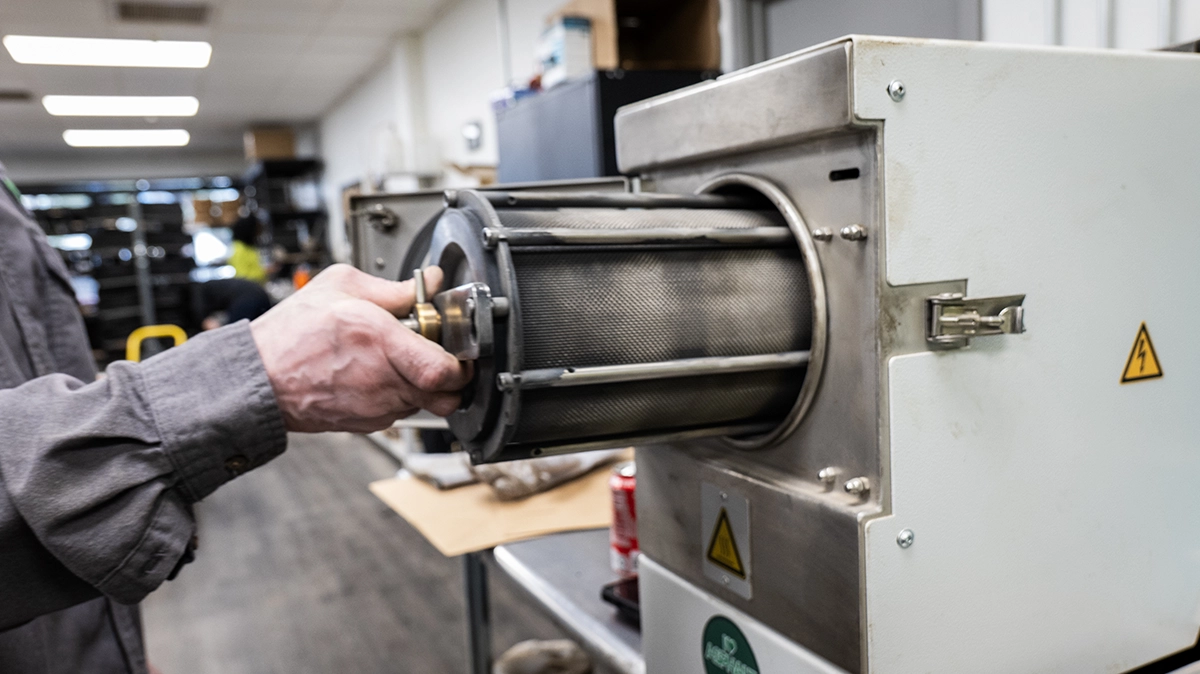 A person slides a cylindrical metal component into a lab furnace or industrial oven inside a workspace.