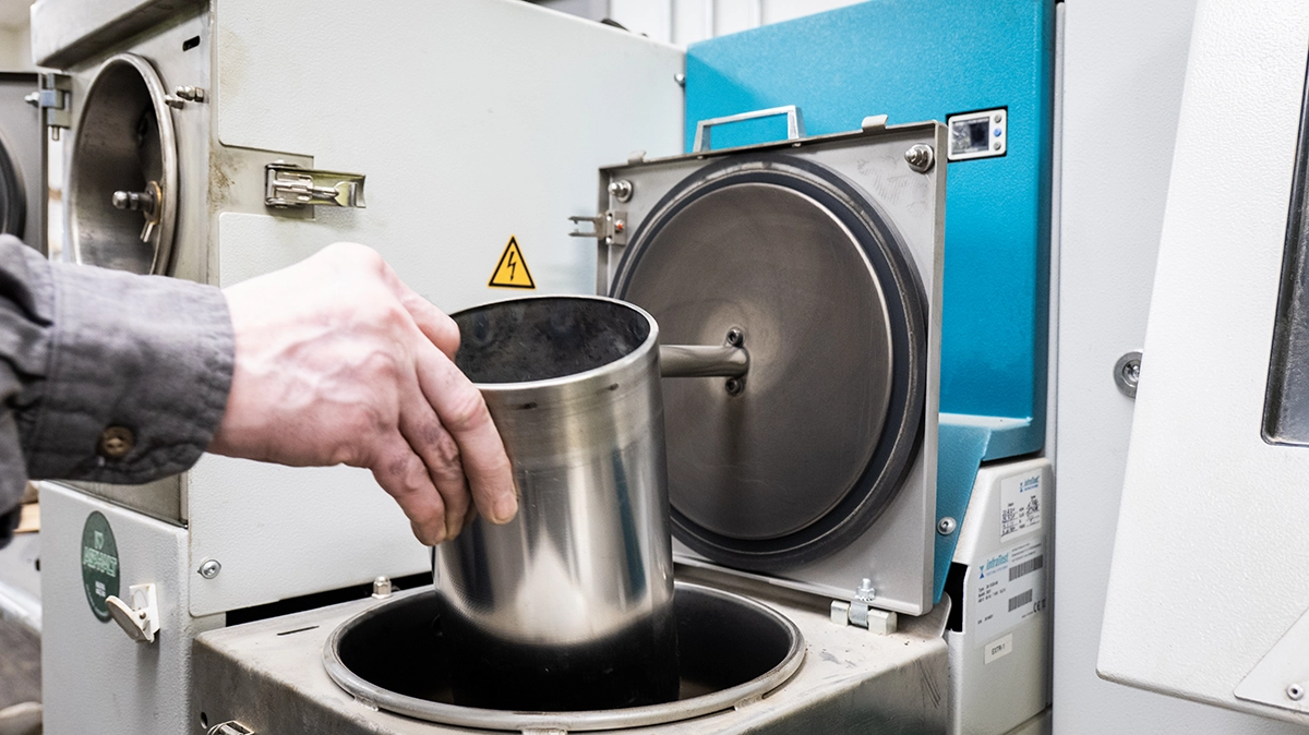 A person places a metal container into a laboratory centrifuge machine with its lid open, preparing for a scientific or industrial process.