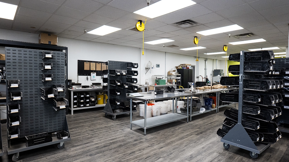 A clean, organized workshop with metal shelves on wheels, worktables, storage bins, and various tools arranged along the walls under bright overhead lighting.
