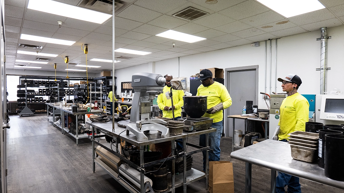 Three workers in yellow shirts stand in an industrial workshop with shelves of equipment, large machines, and workbenches visible around them.