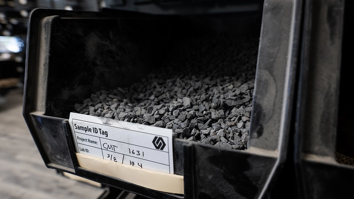 A black storage bin filled with small, dark grey gravel pieces, labeled with a Sample ID Tag showing project name, lab ID, and a logo.