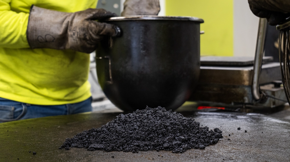 A person wearing gloves stands behind a metal container, with a pile of black granular material on a nearby surface.