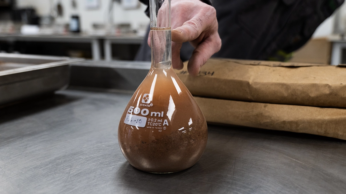 A person holds a 500 ml Erlenmeyer flask filled with a brown liquid and sediment on a metal table, with paper bags and laboratory equipment in the background.