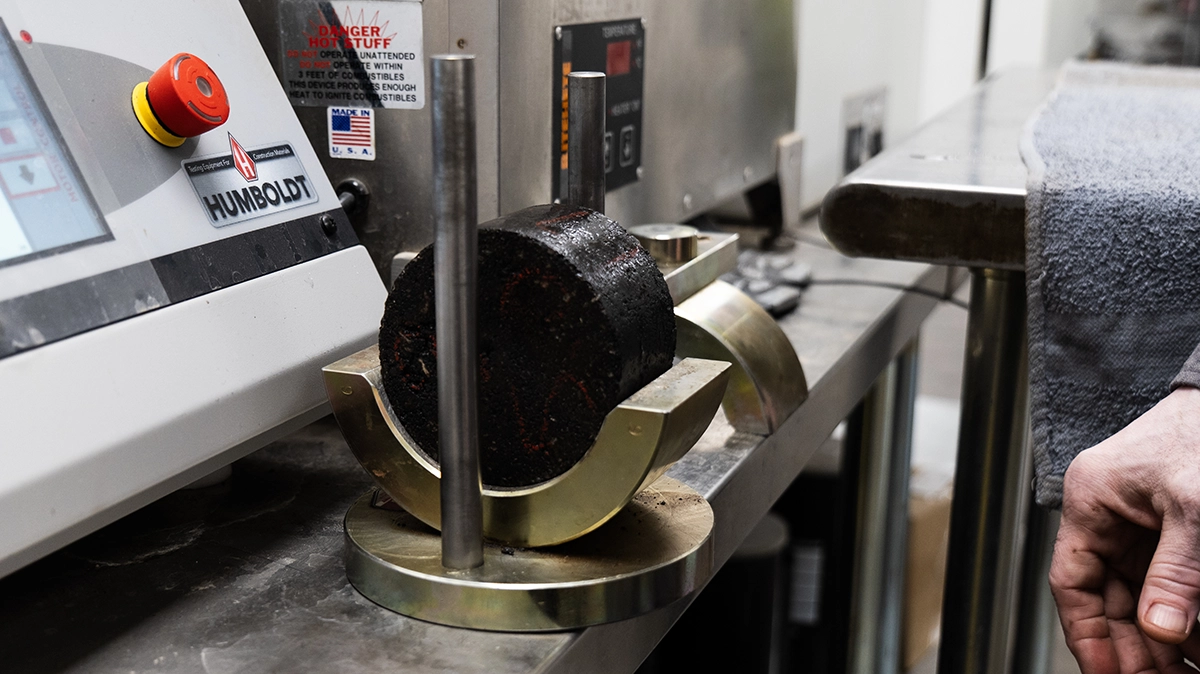 A cylindrical asphalt core sample is positioned on a metal holder next to laboratory equipment, with a person's hand visible on the right.