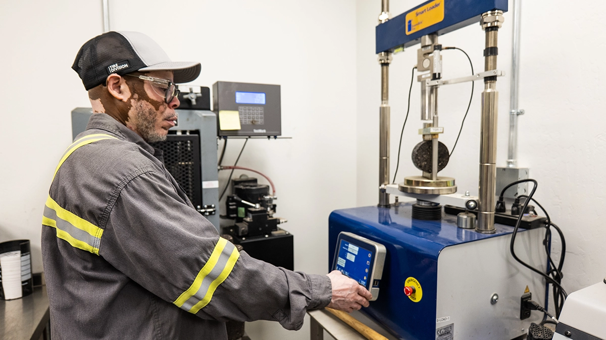A man in safety glasses and workwear operates a laboratory testing machine with a digital interface in an industrial lab setting.