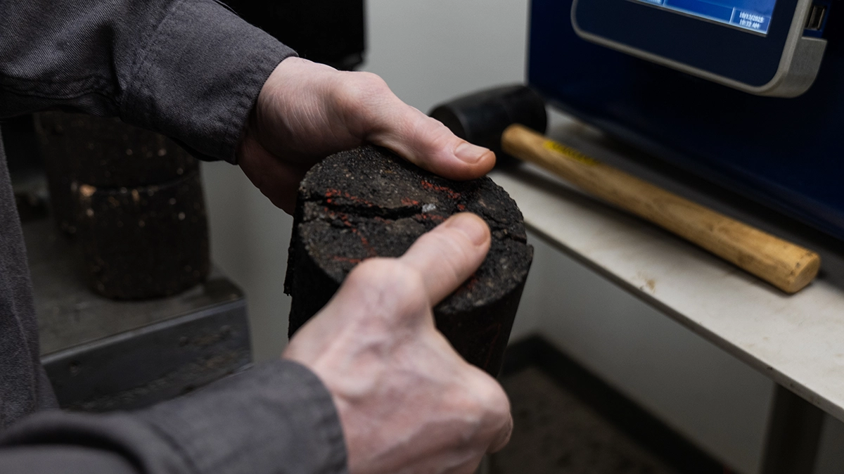 A person’s hands are breaking a cylindrical asphalt sample in half on a table, with a hammer and lab equipment nearby.