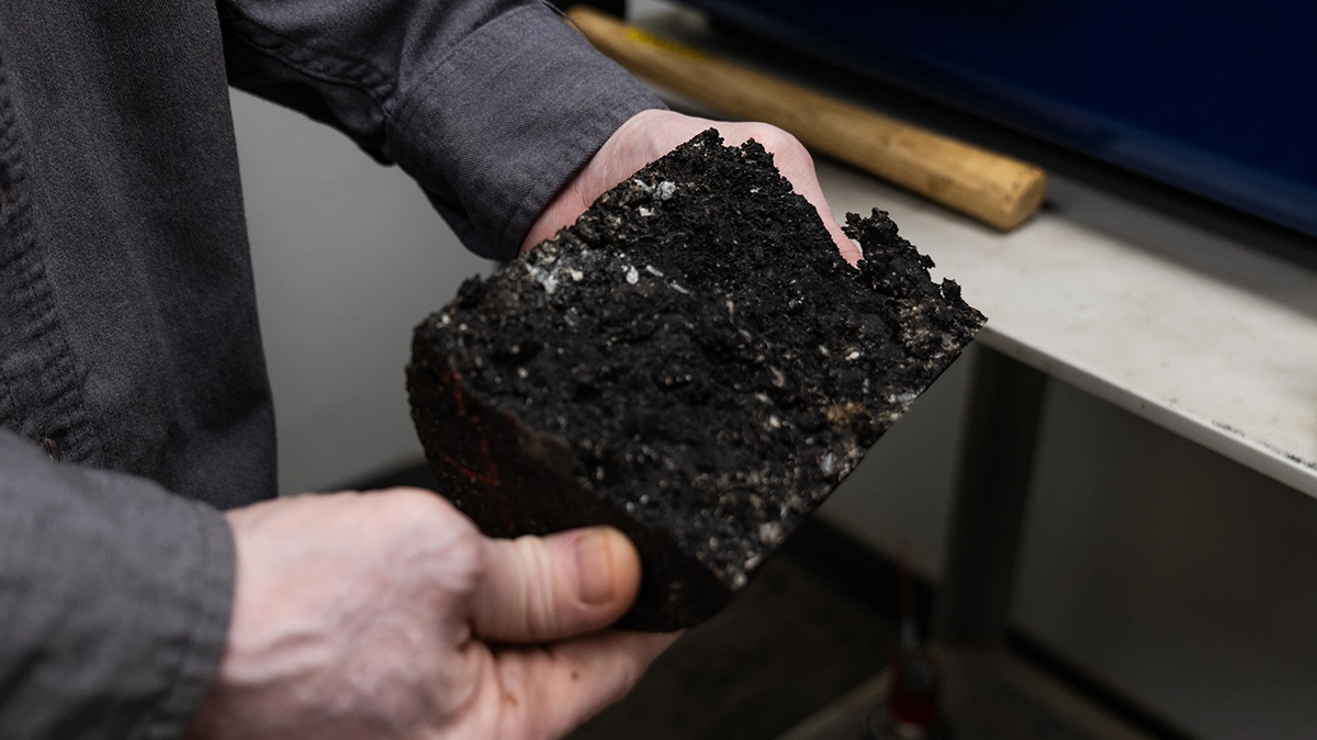 A person holds a large, dark, rectangular block of compacted soil or asphalt in both hands over a table with a wooden tool visible in the background.
