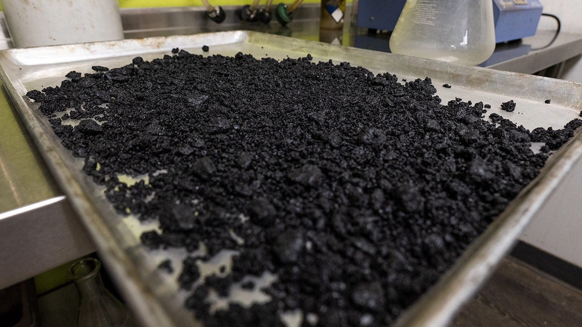 A metal tray filled with a loose, coarse black granular substance sits on a stainless steel countertop in a laboratory setting.