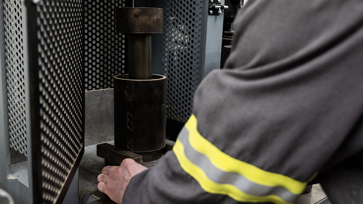 A person operates a machine in an industrial setting, with one hand adjusting a metal component and wearing a uniform with reflective stripes.