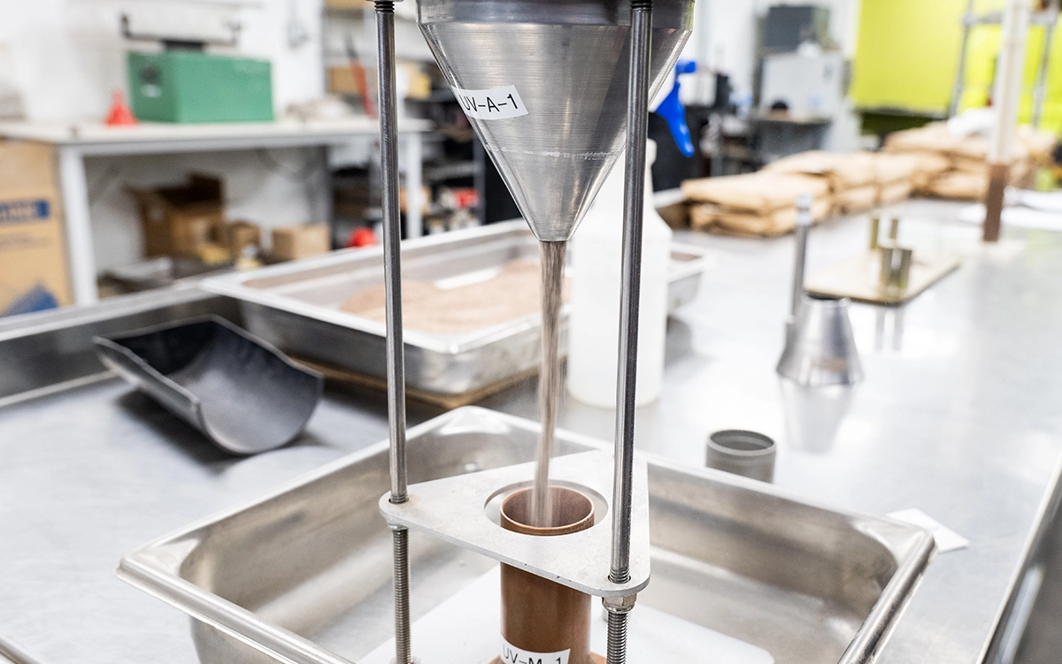 A metal funnel dispenses fine powder into a cylindrical container on a laboratory workbench, with trays and equipment visible in the background.
