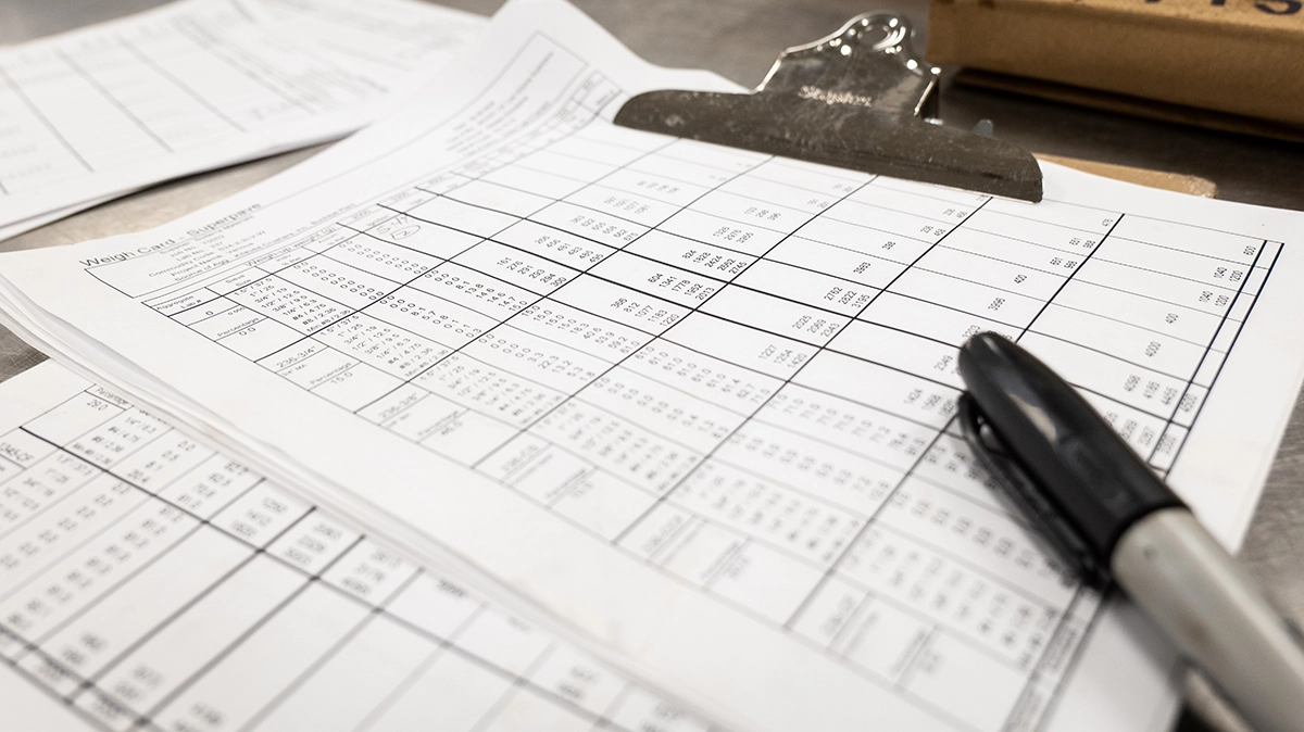 A close-up of a clipboard holding printed tables and data sheets, with a black marker resting on top.