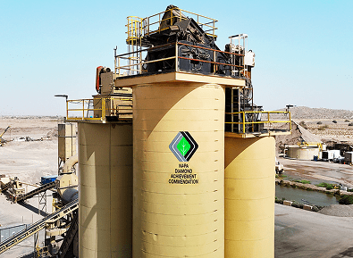 Large industrial silos with yellow exteriors and railings, displaying the "NAMA Diamond Achievement Commendation" logo, situated in an arid, open landscape.