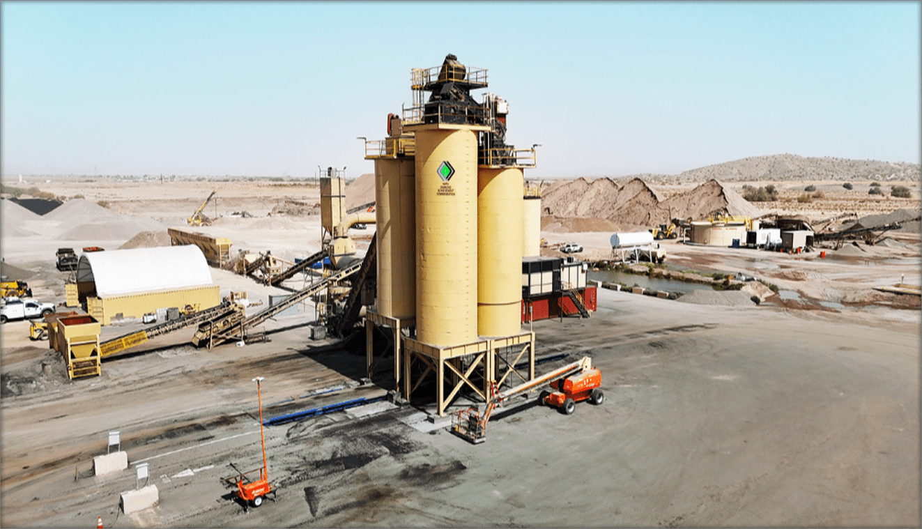 A large yellow industrial facility with silos and construction equipment is set in a dry, open landscape with piles of gravel and dirt in the background.